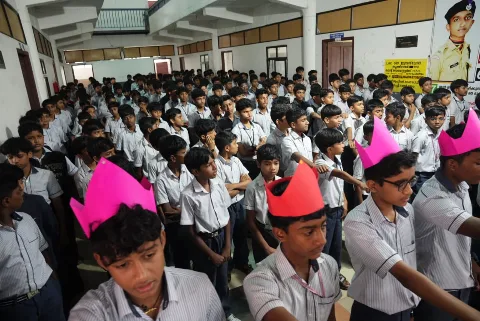 Rows of students in school uniform packed closely together, two in the foreground wearing pink and red paper crowns