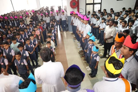 Rows of students facing each other along a hall, young children in navy vests on one side and students in coloured paper crowns on the other