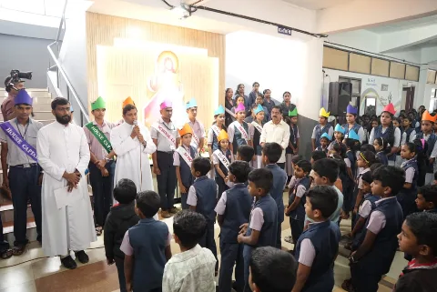 Students in paper crowns and sashes in front of a lit framed religious image while a man in white speaks into a microphone
