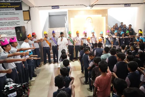 Students with their arms stretched forward while a man in white speaks into a microphone, with a video camera at the edge of the picture